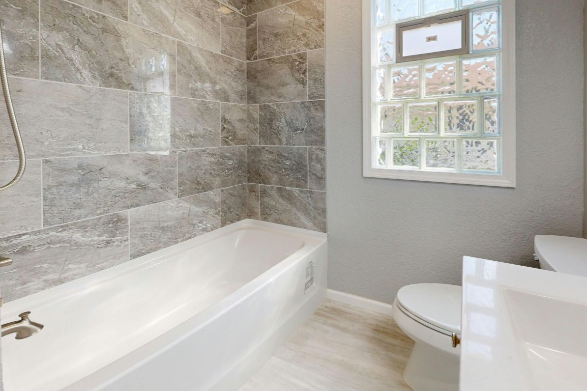 Design ohne Titel – 2 Sanitär und Badmodernisierung der Kösters Sanitär und Heizung GmbH & Co. KG aus Mühlheim an der Ruhr mit Einblick in ein modernes Badezimmer mit großen Fließen, Badewanne, WC, Waschbecken und Fenster mit Glasbausteinen.
